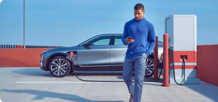 Man walking holding his phone outdoors, with a stationary gray EV connected to the charging station