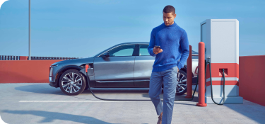 Man walking holding his phone outdoors, with a stationary gray EV connected to the charging station